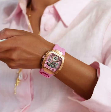 Woman wearing a pink luxury watch with chronograph subdials and a rose gold case, holding a cup and saucer, dressed in a light pink button-up shirt.