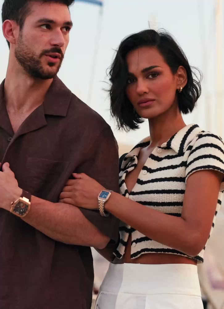 A man and woman stand at a marina with sailboats in the background, both wearing stylish wristwatches; the man in a brown shirt and the woman in a black and white striped top and white pants.