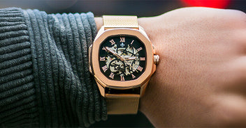 Wrist close-up of a gold-toned watch with a mesh bracelet featuring a black dial, Roman numeral hour markers, and visible skeletal movement details under sapphire-coated crystal.