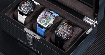 Three men's luxury watches with chronograph subdials and sapphire-coated crystals displayed in a black watch case with a transparent lid on a dark textured surface.
