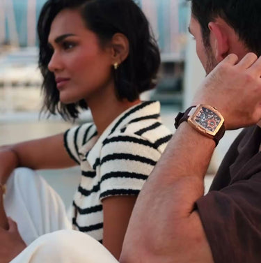 Couple seated beside a marina with boats in the background a man wearing a gold square watch and a woman in a striped top beside him