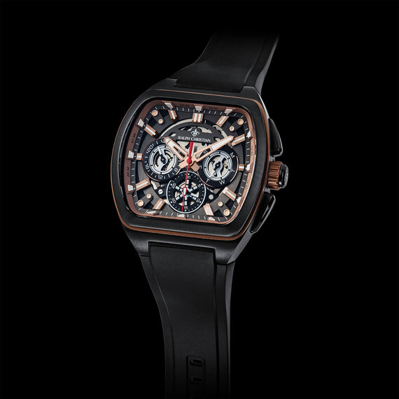 A luxury chronograph watch with a black rubber strap, a matte black case, and a detailed skeleton dial featuring rose gold-toned hands and markers, set against a solid black background.