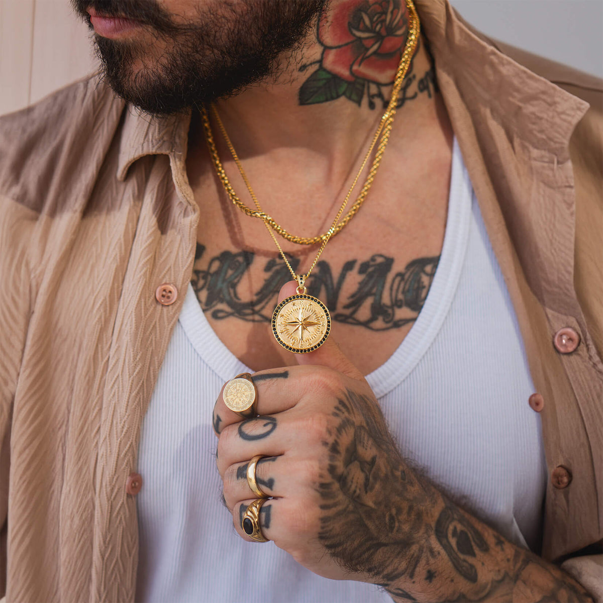 A man wearing layered gold chains featuring The Gold Compass Pendant - Essentials Collection, paired with multiple gold rings, a white tank top, and a beige open shirt.