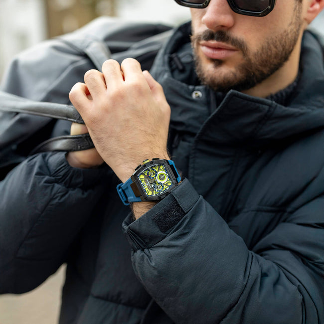 Man wearing a black jacket and sunglasses, showcasing a bold men's watch with a square black and blue case, neon green and black dial, and chronograph subdials on his wrist.