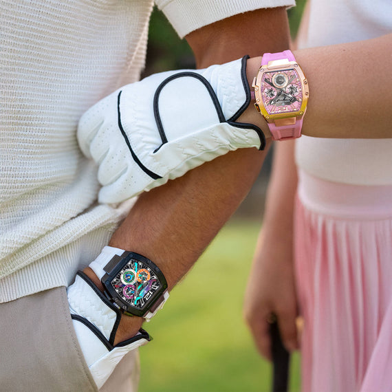 Two women wearing stylish watches outdoors. One watch has a pink strap and gold case with a floral screen, while the other has a black strap and case with a colorful screen.