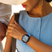 A woman wearing a wide-brimmed hat and blue sleeveless top, showcasing The Intrepid Sport Ladies - Blue Mirage watch with a bold black and blue case and strap, featuring a detailed dial with day and date functions, embodying a statement luxury watch for women. Bold by Design.