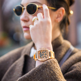 A woman wearing sunglasses and a brown coat showcases The Intrepid Sport Ladies - Sunkiss Bronze watch with a bold bronze case, visible chronograph subdials, and an orange strap.
