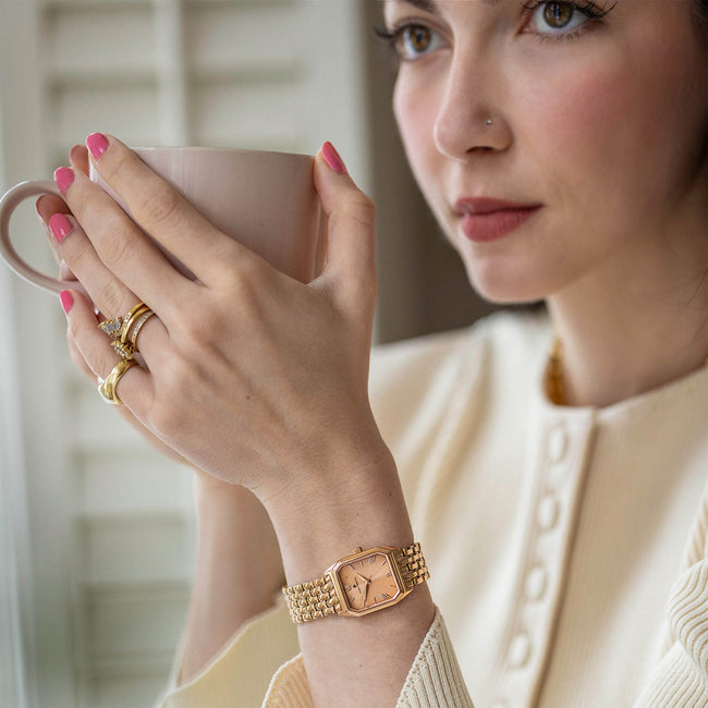 A close-up lifestyle shot of a woman wearing The Valencia - Rose Allure, a rose gold women's luxury watch with a rectangular dial and link bracelet, holding a beige mug.