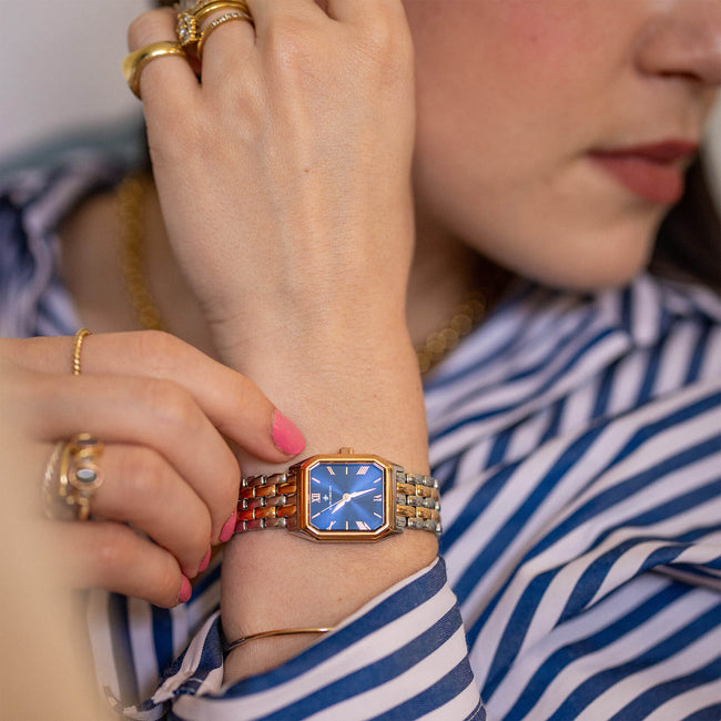 A close-up lifestyle shot of a female wrist wearing The Valencia - Twilight Bloom, a bold women's luxury watch with a square rose gold case, blue dial, and metal bracelet, styled with gold rings and a striped shirt.
