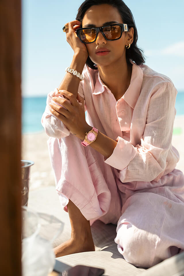 Woman sitting outdoors on a sunny day wearing oversized brown sunglasses, a light pink linen shirt and pants, and a rose gold watch with a matching bracelet on her wrist.