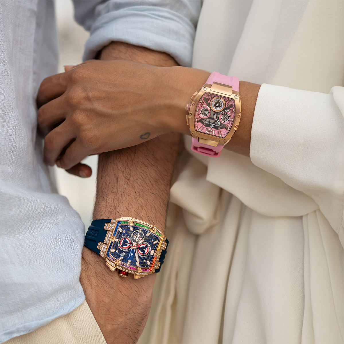 Close-up of two wrists side by side wearing bold luxury watches; one with a pink strap and rose gold case, the other with a dark blue strap, gold case, and colorful dial accents.