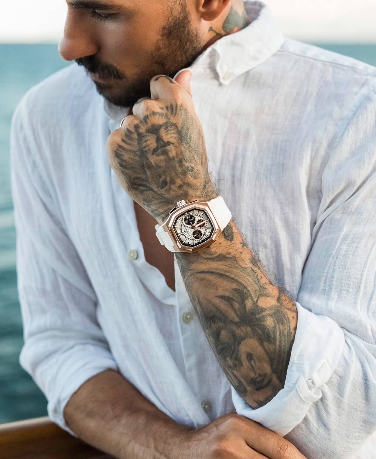 Man wearing a white shirt with a tattooed arm, holding a watch.