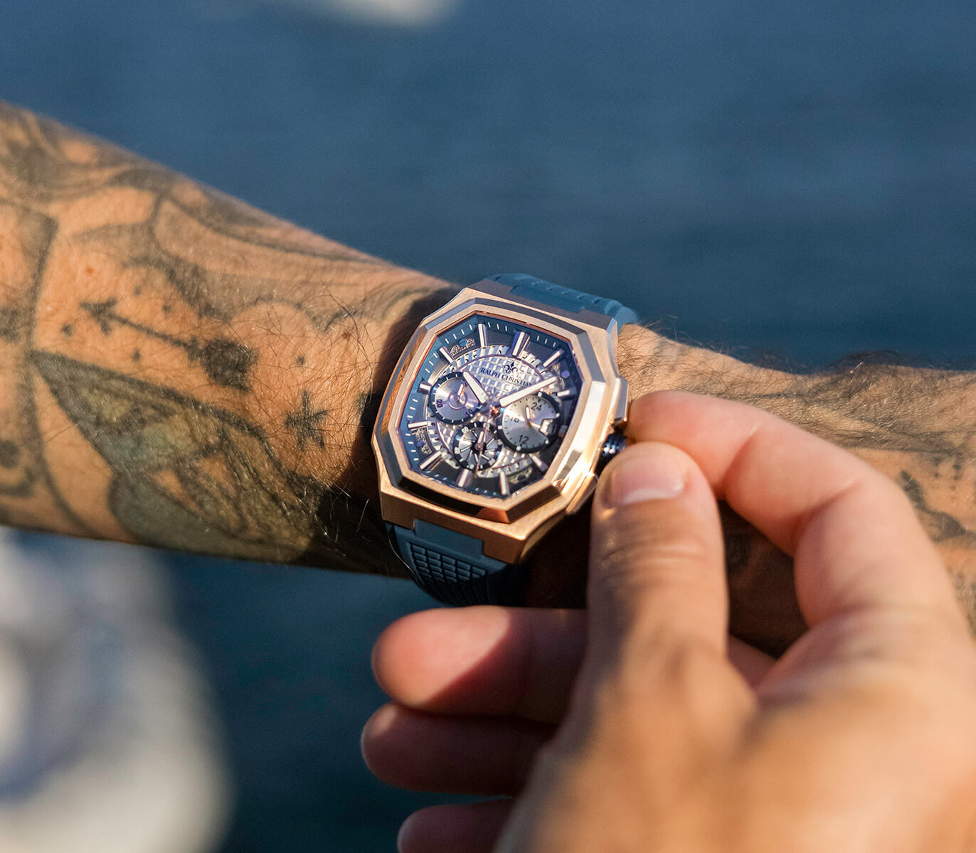 Person wearing a detailed wristwatch with a tattooed arm, water in the background