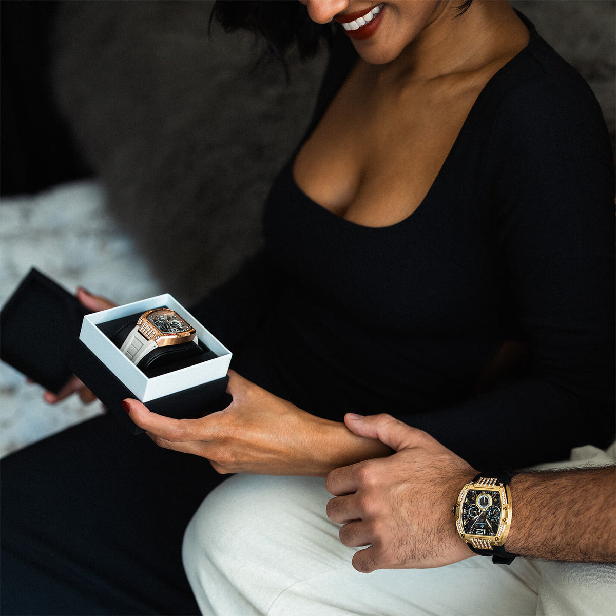 A woman holds an open box containing a gold and black watch while a man wears a matching ornate watch on his wrist.