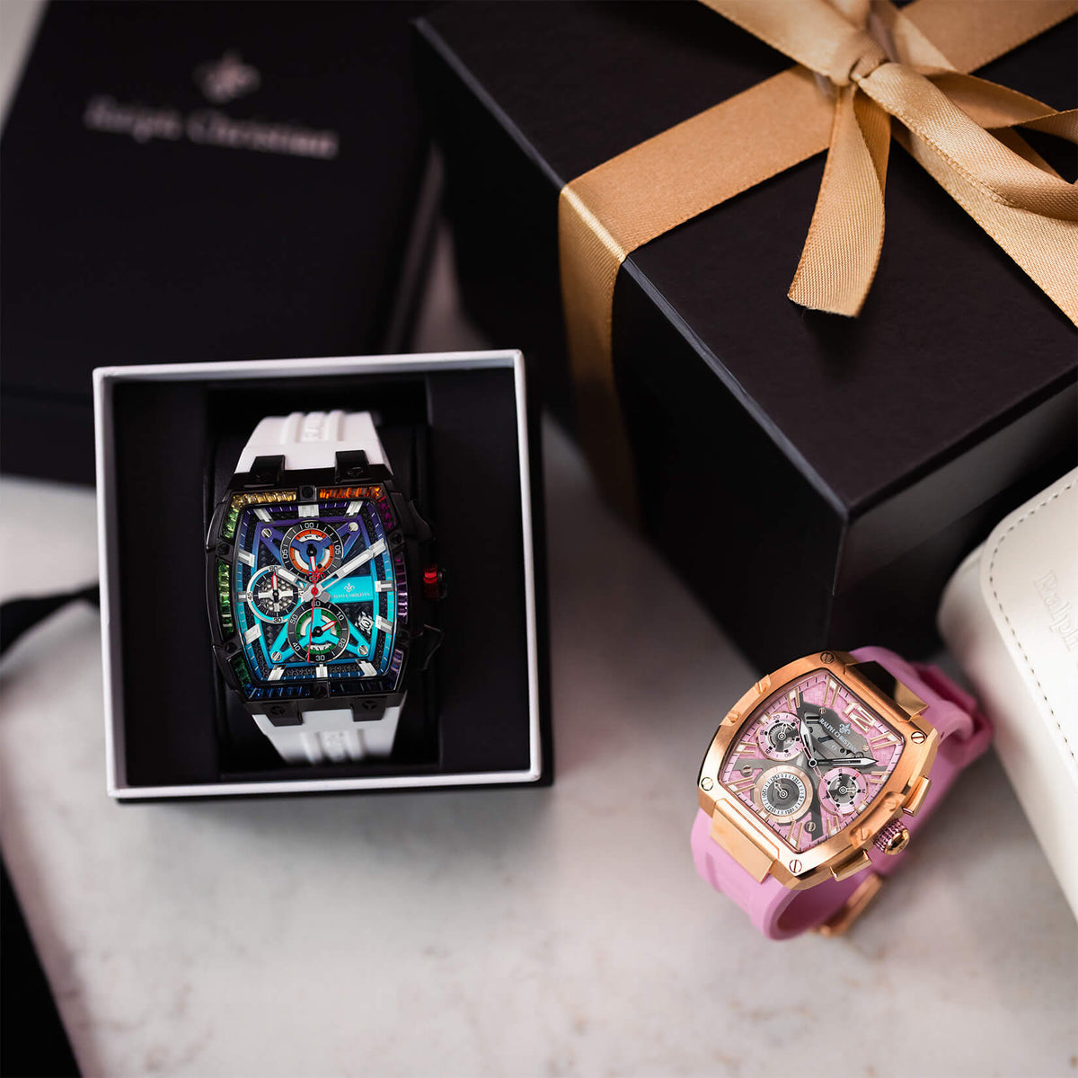 Two statement watches displayed with gift boxes one featuring a black and white case with colorful chronograph subdials and another with a rose gold case and pink strap