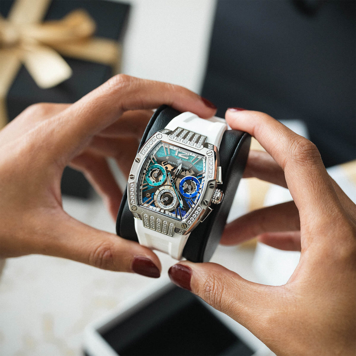 Hands holding a statement watch with a white strap, silver-toned case, blue chronograph subdials, and a crystal accented bezel over a gift box background