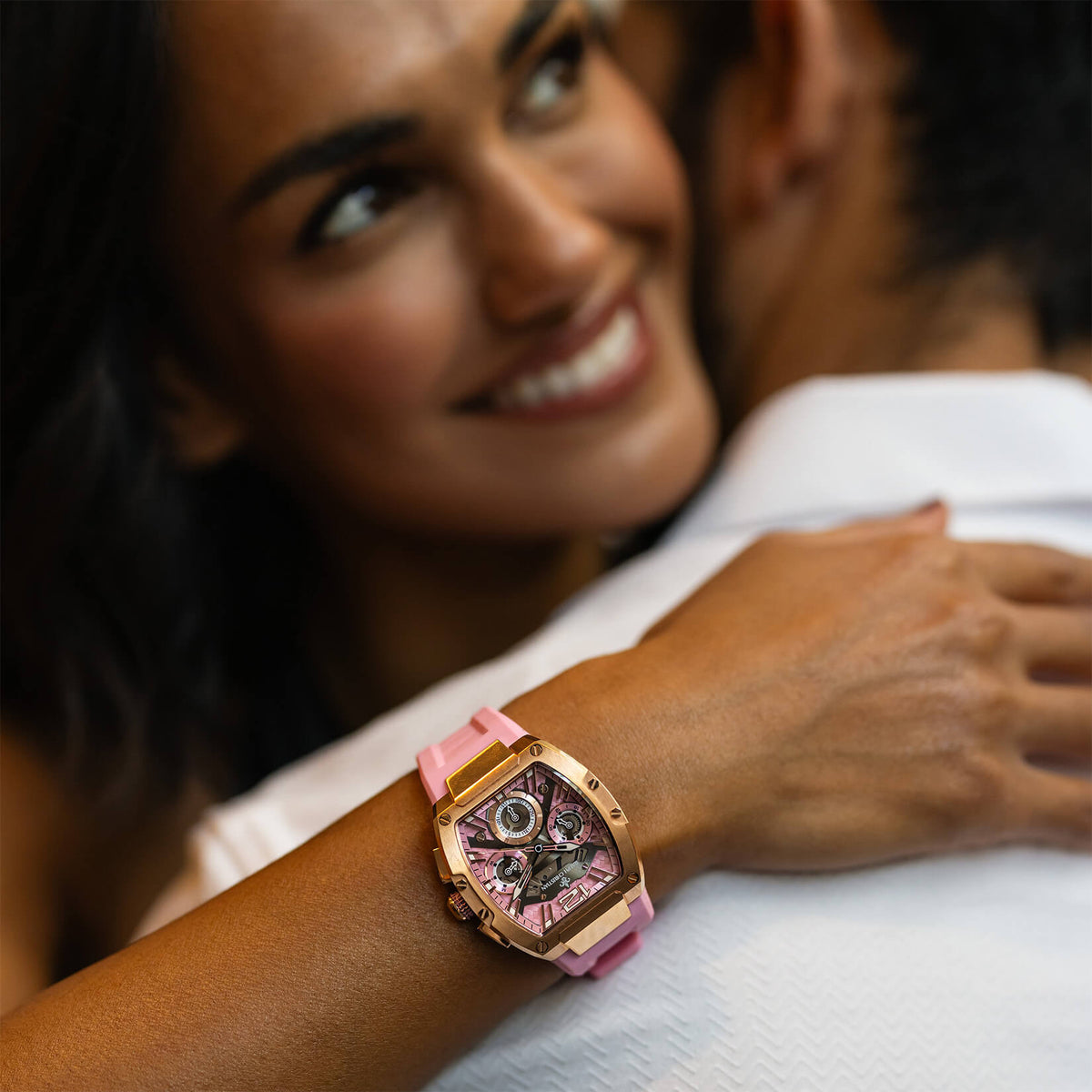 Smiling woman embracing a man while wearing a rose gold watch with a pink strap and intricate dark dial on her wrist