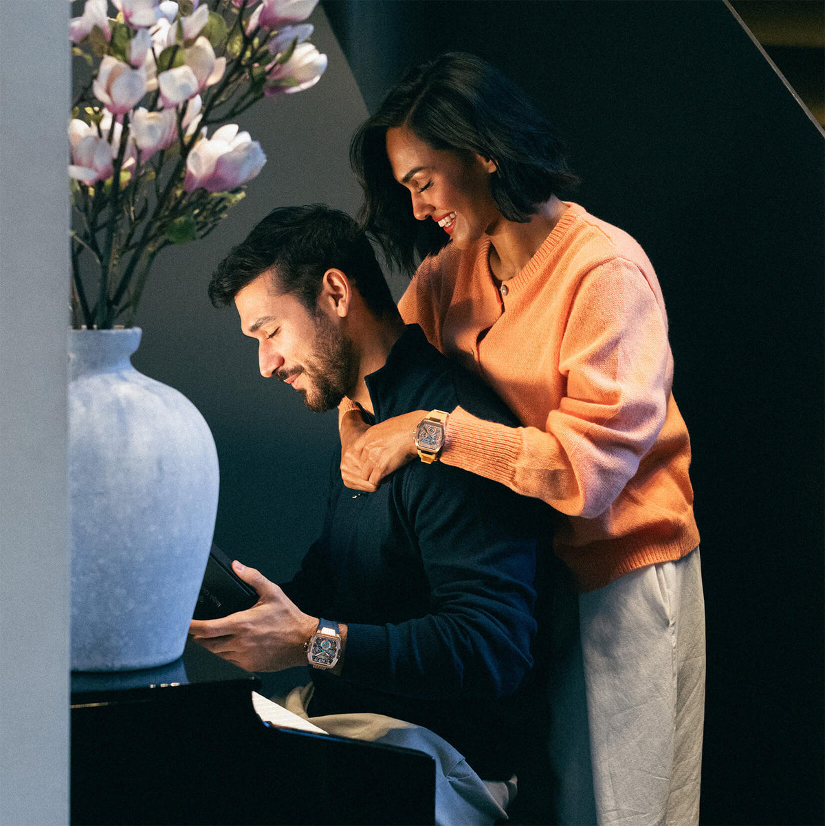 Couple sharing a warm moment beside a piano, both wearing luxury watches, with a vase of pink flowers in the background and soft natural lighting