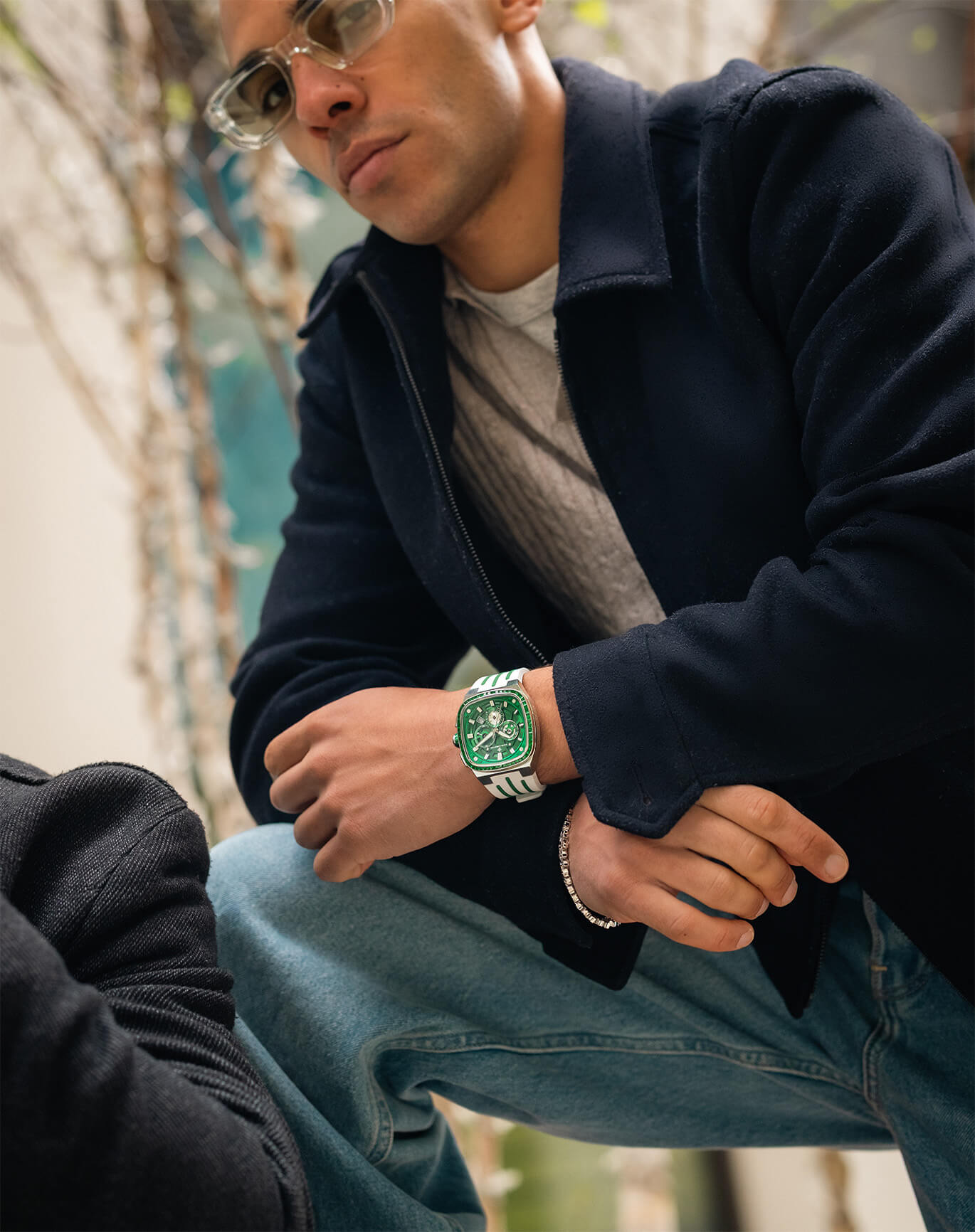 Man wearing clear glasses, a navy jacket, and jeans, displaying a bold green watch with a chronograph dial and striped strap on his wrist in an outdoor setting.