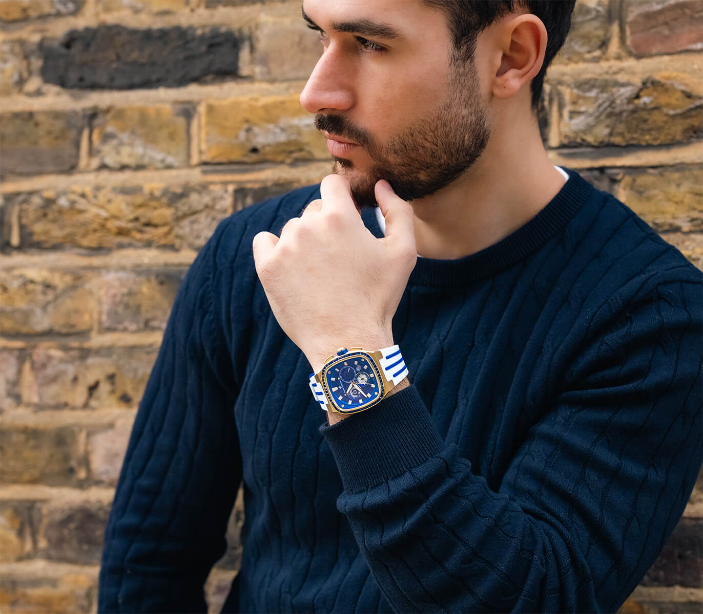 Man wearing a navy blue cable-knit sweater and a bold gold and blue chronograph watch with a white and blue striped strap, posing thoughtfully against a brick wall.
