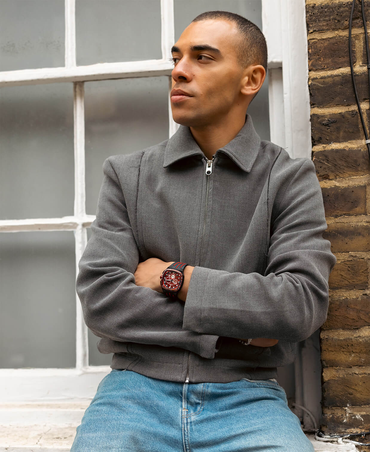 Man wearing a bold timepiece with a red dial and brown leather strap, styled with a gray zip-up jacket and light blue jeans while leaning against a brick wall near a window.