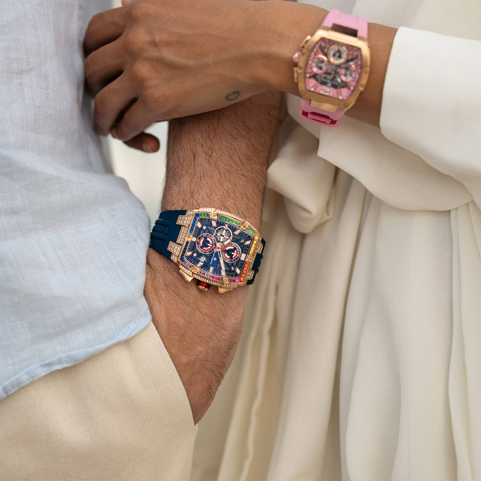 Two wrists crossed showing bold luxury chronograph watches with colorful cubic zirconia iced bezels, one with a blue strap and blue dial, the other with a pink strap and pink dial, both paired with casual and elegant light-colored clothing.