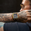 Man with tattoos wearing a gold and blue watch on a dark background