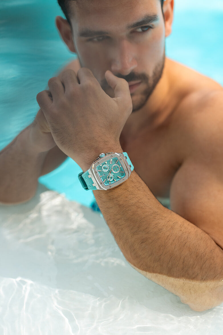 Close-up lifestyle shot of a man in a pool wearing a men's luxury chronograph watch with a square silver case, aqua dial with subdials, and matching aqua strap.