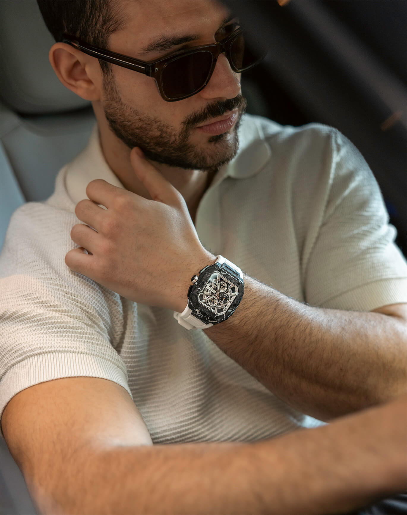 Man wearing dark sunglasses and a white short-sleeve polo shirt seated while adjusting a bold luxury watch with a large square dial and white strap on his wrist.