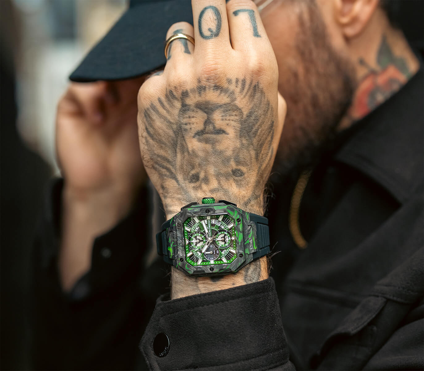 Close-up of a man's wrist wearing a bold green and black chronograph watch with a geometric carbon fiber case, intricate dial, and black strap, styled with visible tattoos.