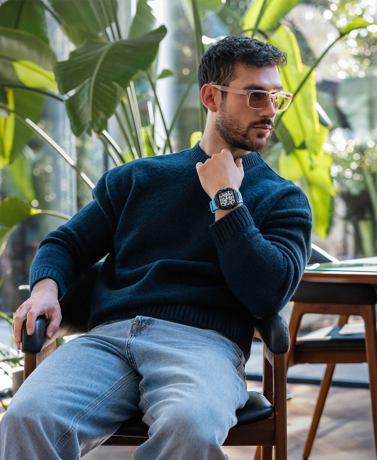 Man wearing light brown square sunglasses and a dark blue sweater sitting indoors on a wooden chair, wearing a bold black watch with a square dial and chronograph subdials on his left wrist.