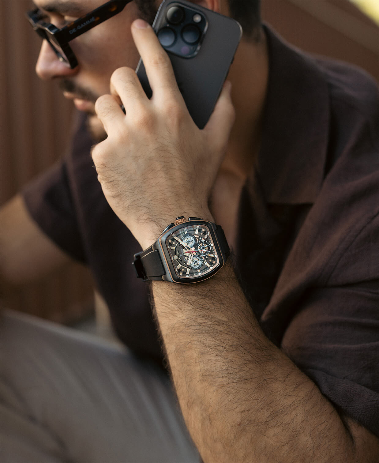 Close-up of a man's wrist wearing a black luxury chronograph watch with multiple subdials, a square case, and a black strap while holding a smartphone to his ear.