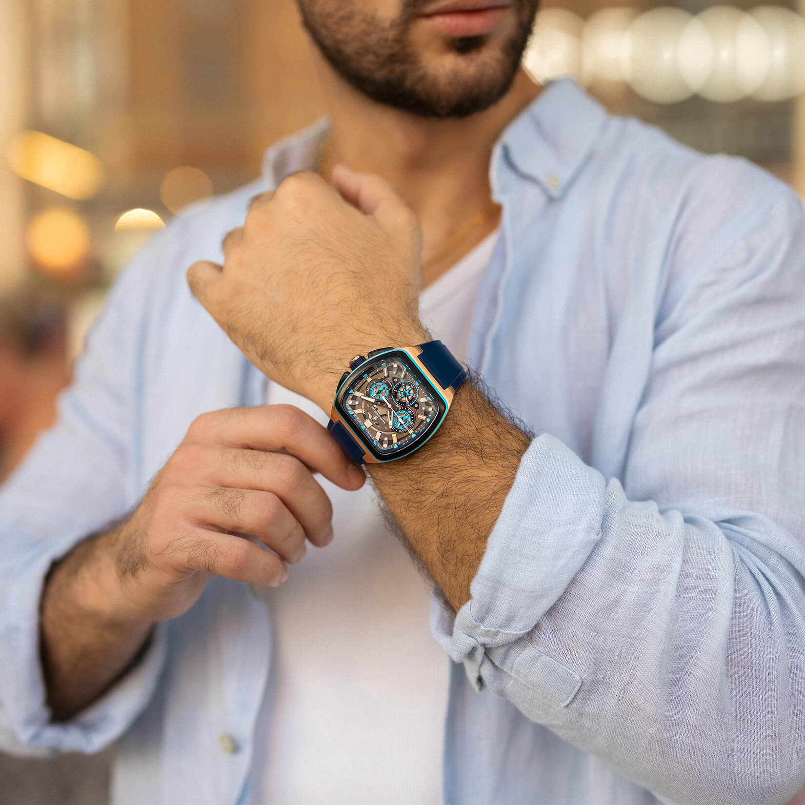 Man wearing a blue-strap wristwatch with a square, transparent dial showcasing intricate mechanical details, paired with a light blue shirt with rolled-up sleeves.