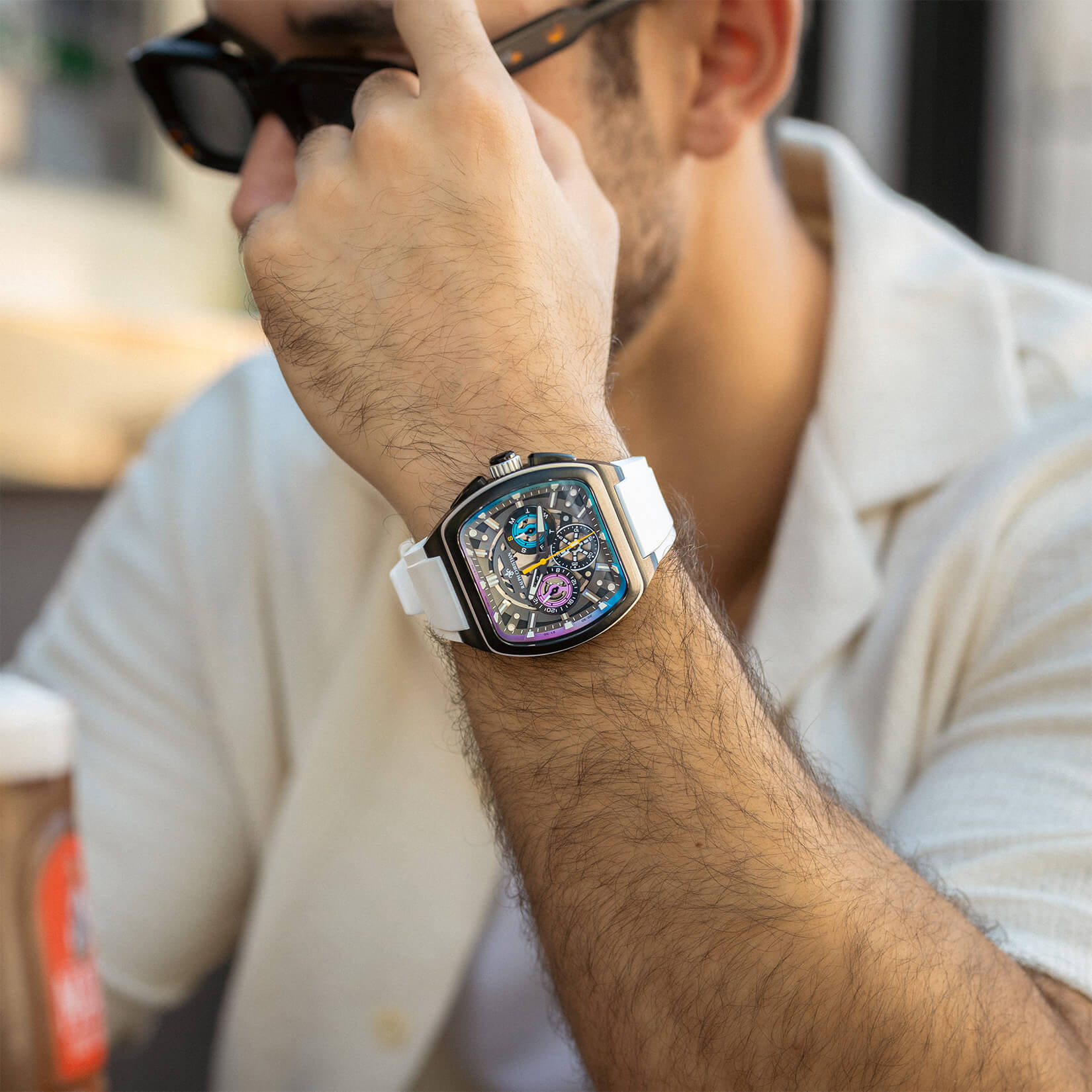 Close-up of a man's wrist wearing a square-shaped luxury watch with a white strap, black bezel, and intricate chronograph subdials visible on the dial, shown in a casual setting.