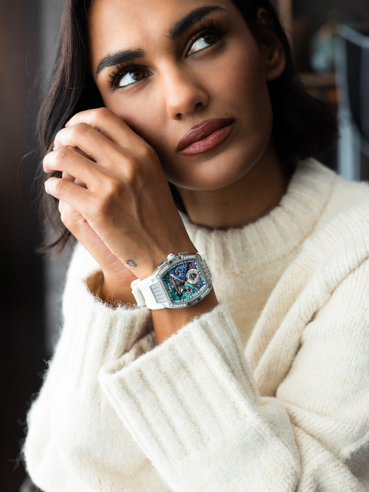 Close up portrait of a woman in a cream knit sweater wearing a silver tonneau watch with a blue dial, crystal details, and a white strap on her wrist